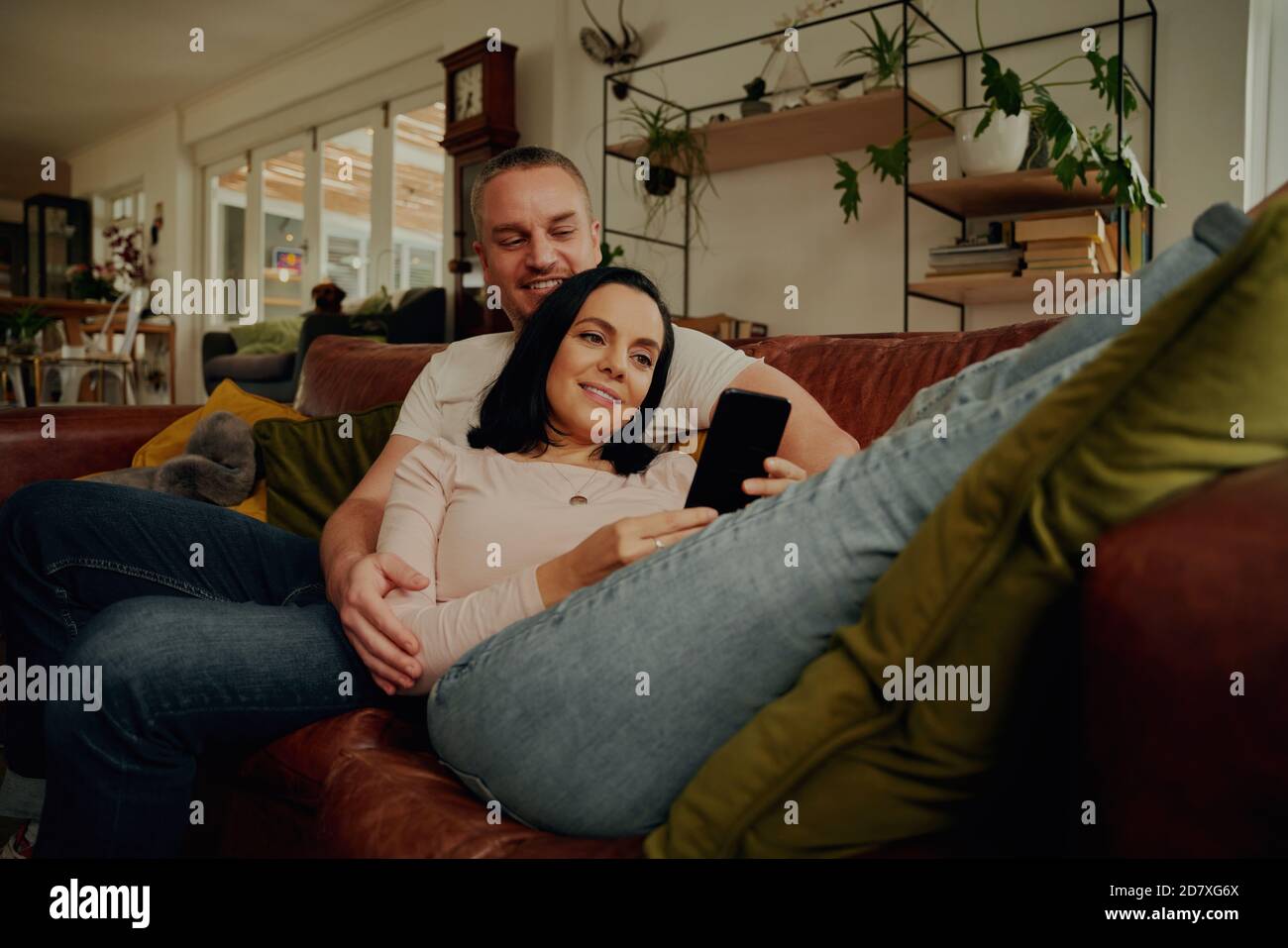 Sorridente giovane donna che usa il telefono mentre si rilassa con il marito divano a casa Foto Stock