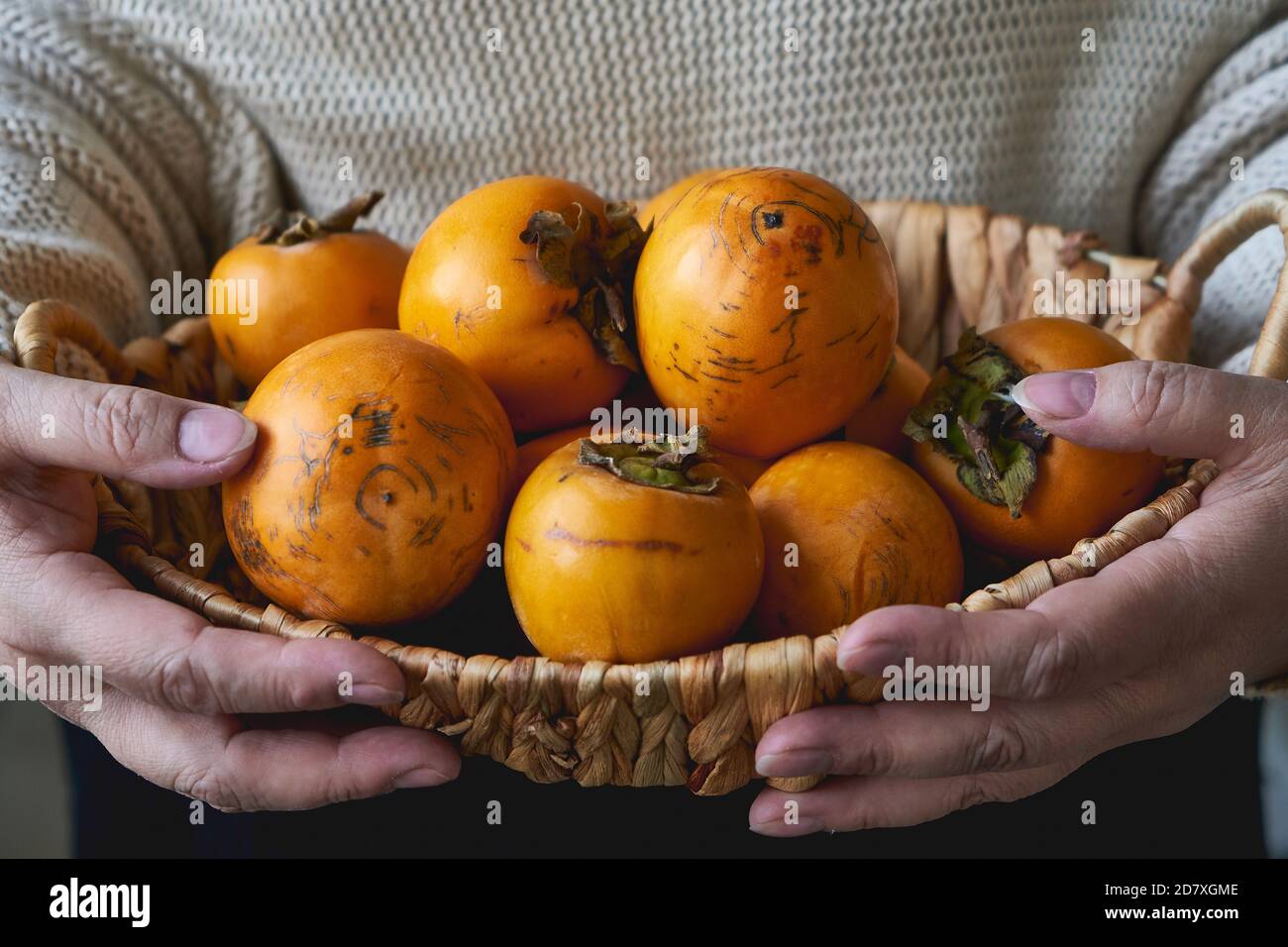 Una donna anziana tiene un cesto con un raccolto di persimmons su un tavolo di legno. Foto Stock