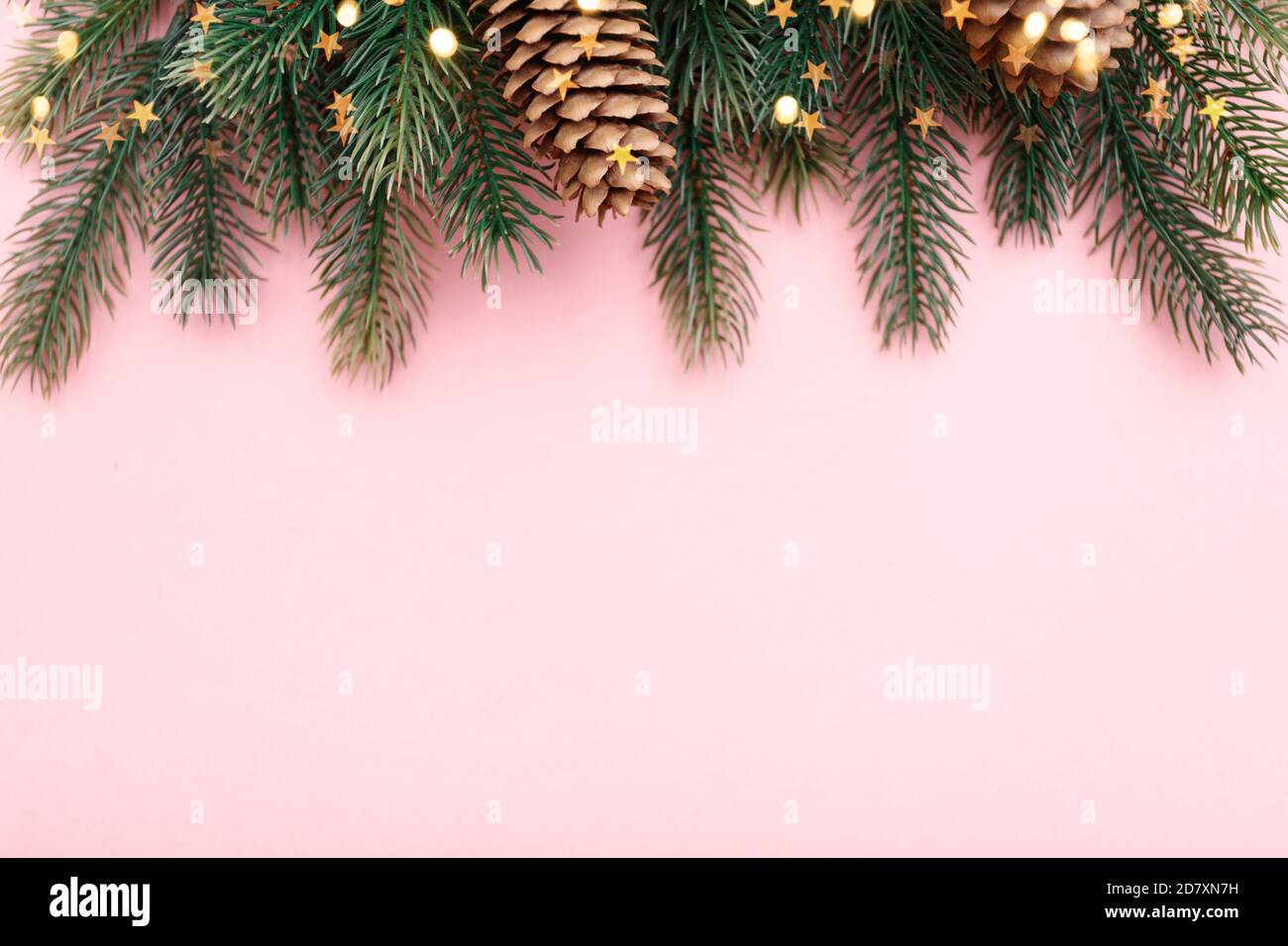Confine natalizio con rami di abete, coni di conifere e luci dorate su sfondo rosa pastello Foto Stock