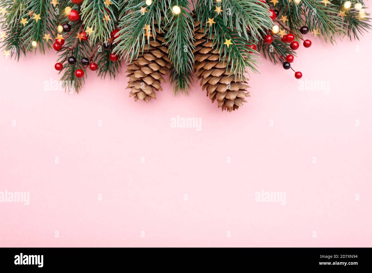 Confine natalizio con rami di abete, coni di conifere, bacche rosse e luci dorate su sfondo rosa pastello Foto Stock