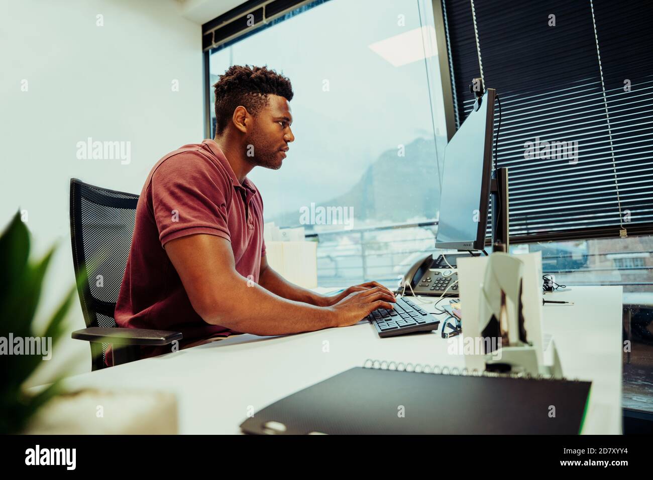 Imprenditore africano americano maschio che cerca i disegni del logo per l'azienda che usa computer portatile desktop in un ufficio elegante Foto Stock