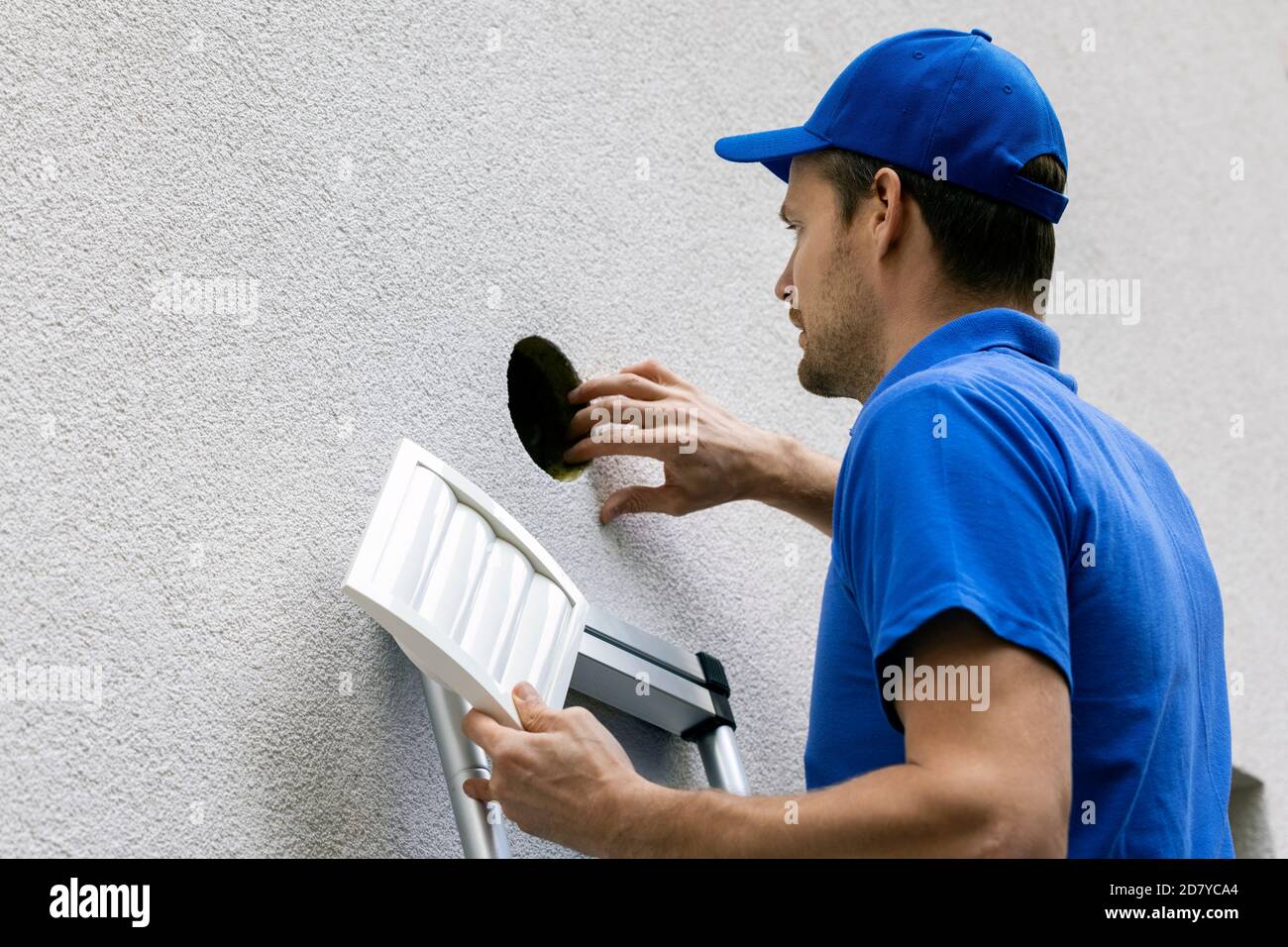 uomo in uniforme blu in piedi su scala all'aperto e fare manutenzione del sistema di ventilazione della casa Foto Stock