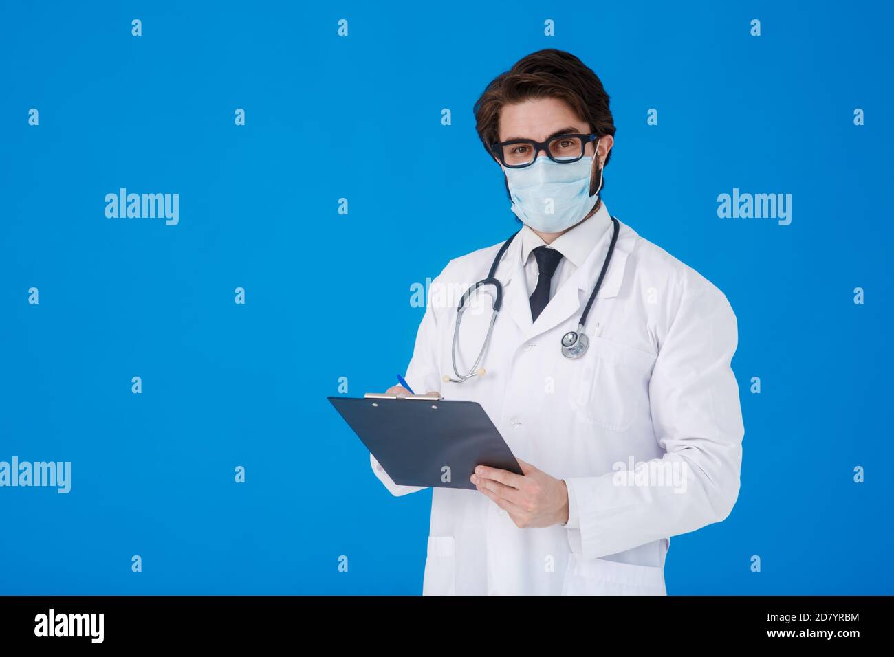 un giovane medico sopportato in un cappotto medico bianco e. una maschera su sfondo blu scrive il paziente reclami su un tablet con documenti e. Foto Stock