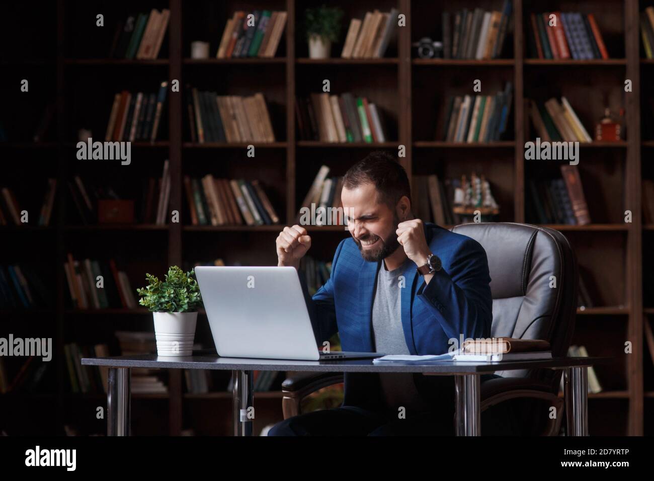 Un giovane uomo d'affari in una tuta blu gioisce del successo e della vittoria guardando lo schermo del portatile. Uomo in biblioteca dell'ufficio Foto Stock