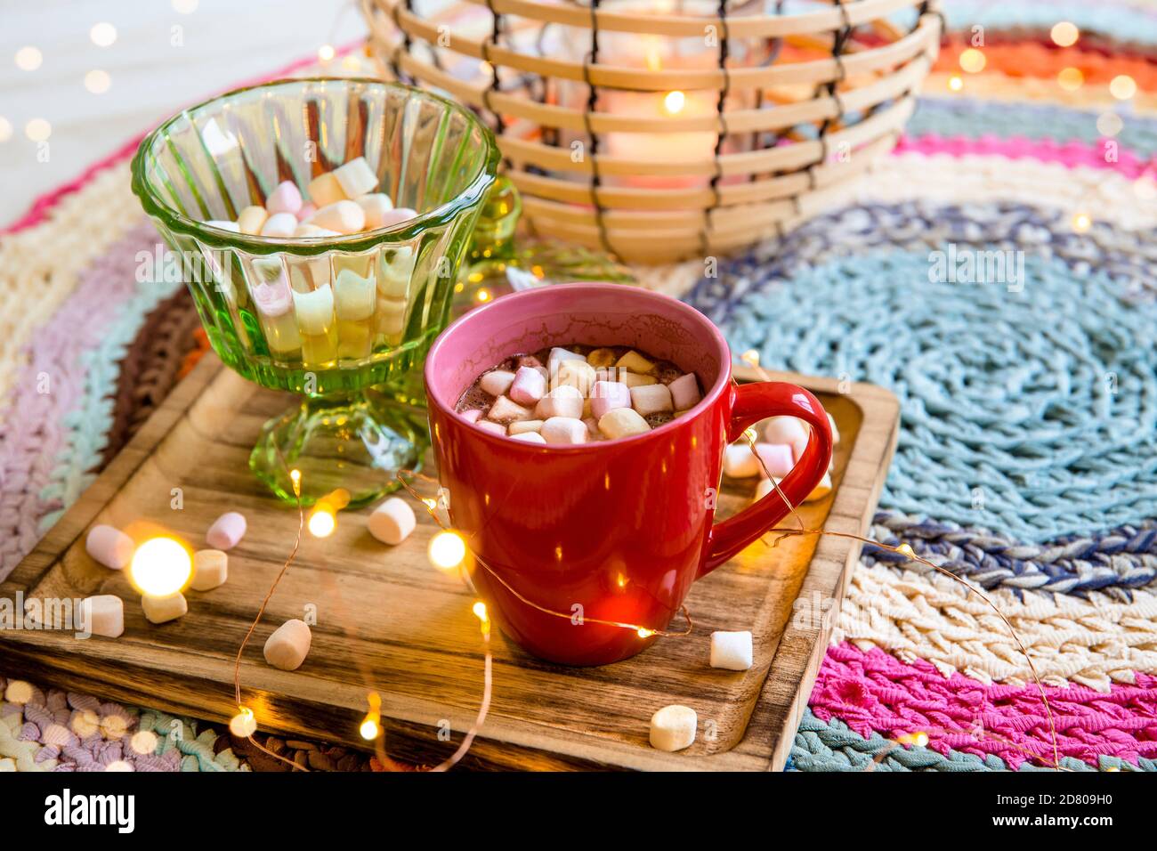 Fuoco selettivo su tazza rossa di cacao con marshmallows di colore pastello, filo metallico nero e lanterna di bambù con candela su tappeto di uncinetto colorato da repur Foto Stock