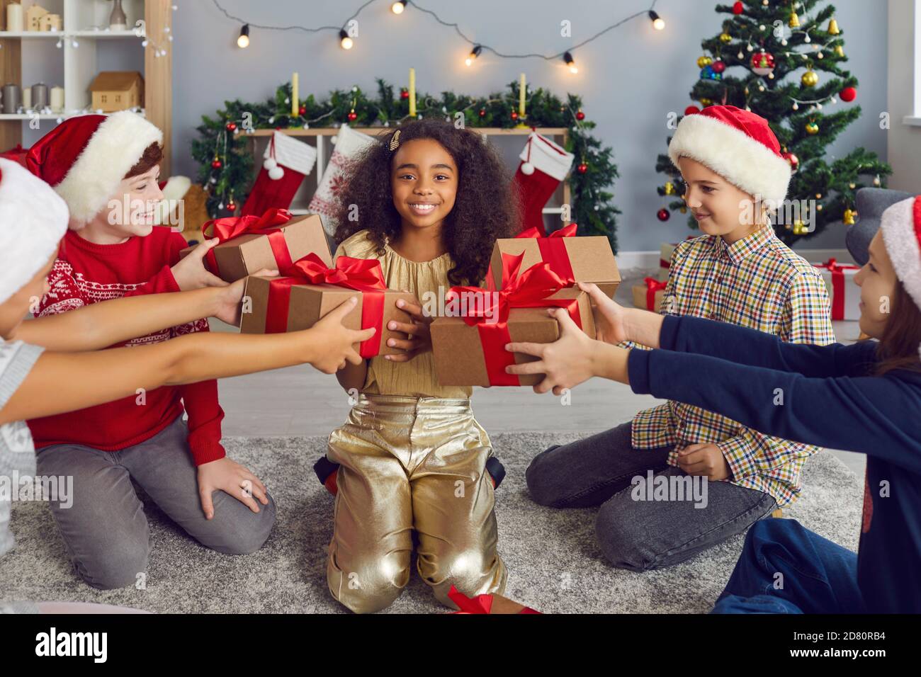 Gruppo di bambini in cappelli di Natale dare doni ad una felice afroamericana ragazza che ha un compleanno. Foto Stock