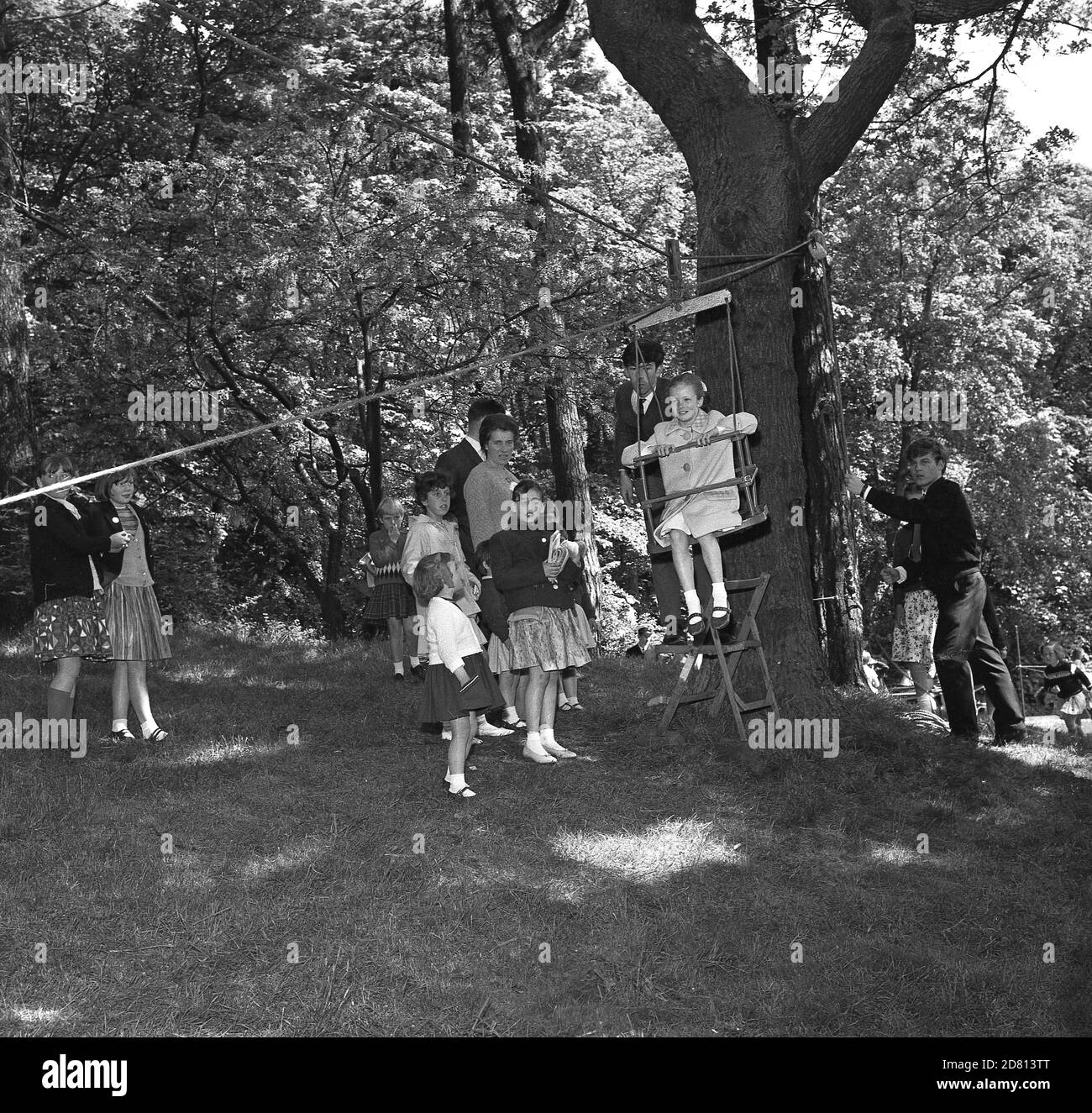 Anni '60, immagine storica di bambini giovani in piedi guardando come una bambina seduta su una sedia swing attaccato a imbracatura e una corda che è legata a un albero, che sta per viaggiare lungo la corda attraverso una zona erbosa, una delle attività in un villaggio estate Fete, Fife, Scozia Foto Stock