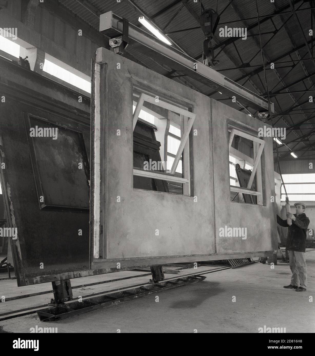 Anni '60, storico, un uomo di lavoro che sposta un pannello esterno in cemento prefabbricato di recente costruzione, con finestre in legno incorporate, che si trova su un rack sospeso appeso al soffitto, Inghilterra, Regno Unito. Foto Stock