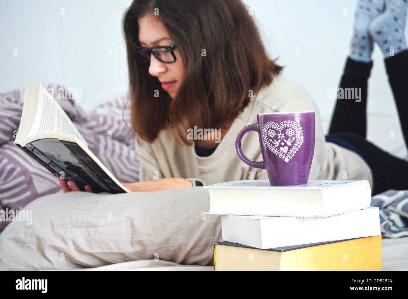 Giovane donna con i capelli corti e gli occhiali marroni in abiti accoglienti leggendo un libro a letto a casa. Mucchio di libri con una tazza di tè decorata viola sulla parte superiore. Foto Stock