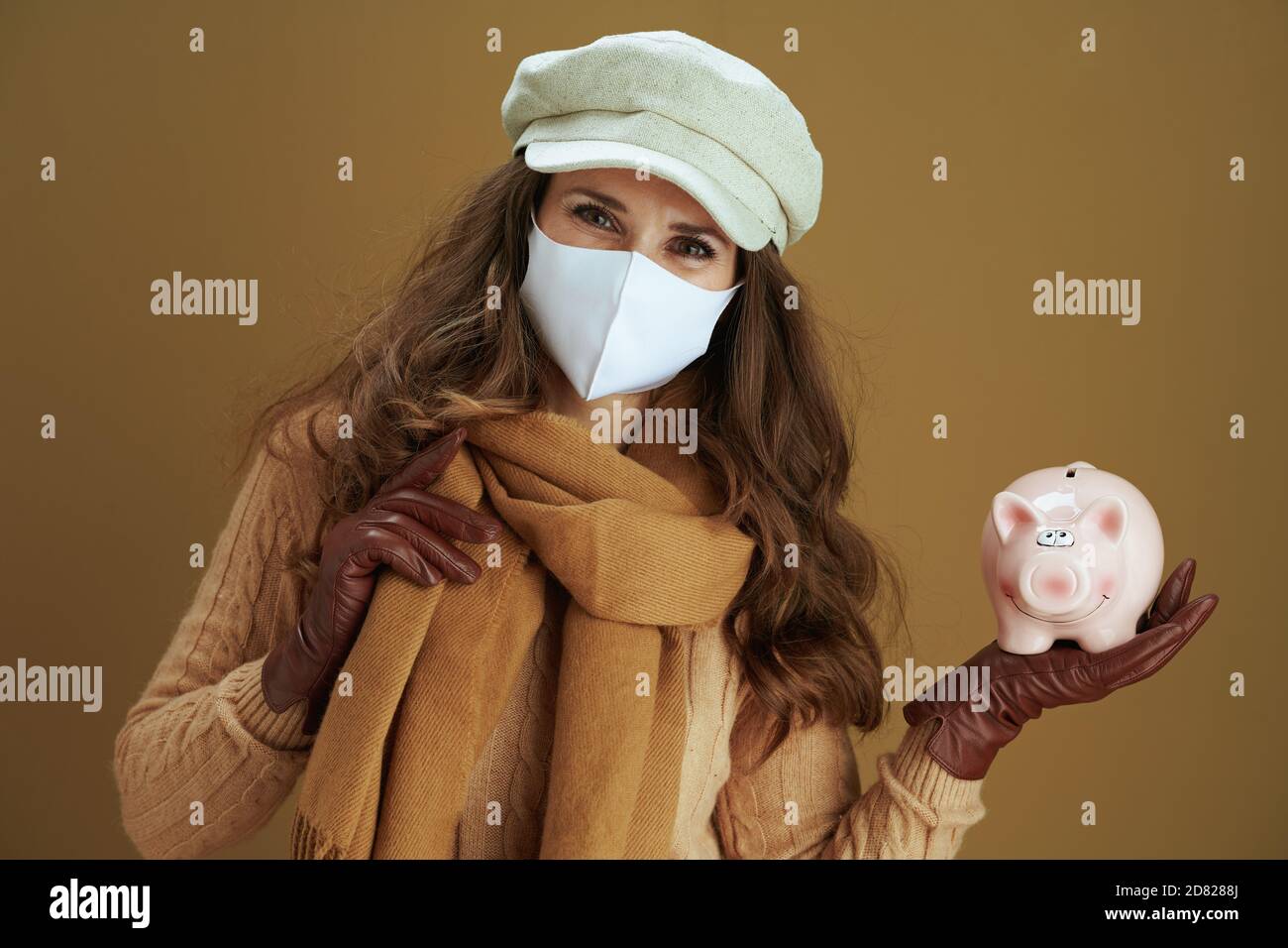 Ciao autunno. Ritratto di donna moderna sorridente in sciarpa con banca piggy e maschera medica bianca su sfondo bronzeo. Foto Stock