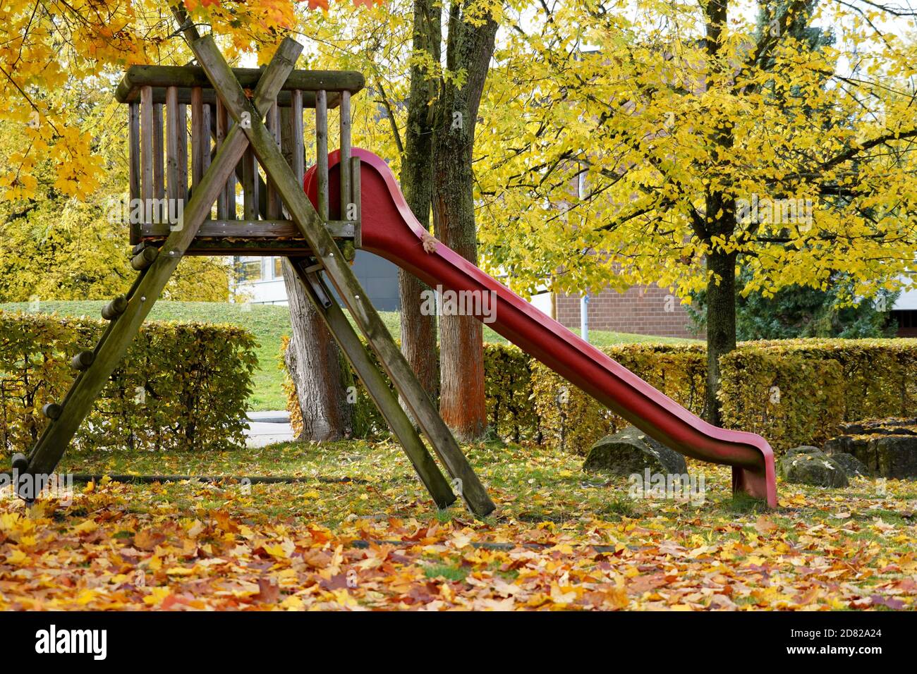 Uno scivolo rosso nel parco in autunno in vista laterale circondato da fogliame in varie sfumature di giallo sia sul terreno che su alcuni alberi. Foto Stock