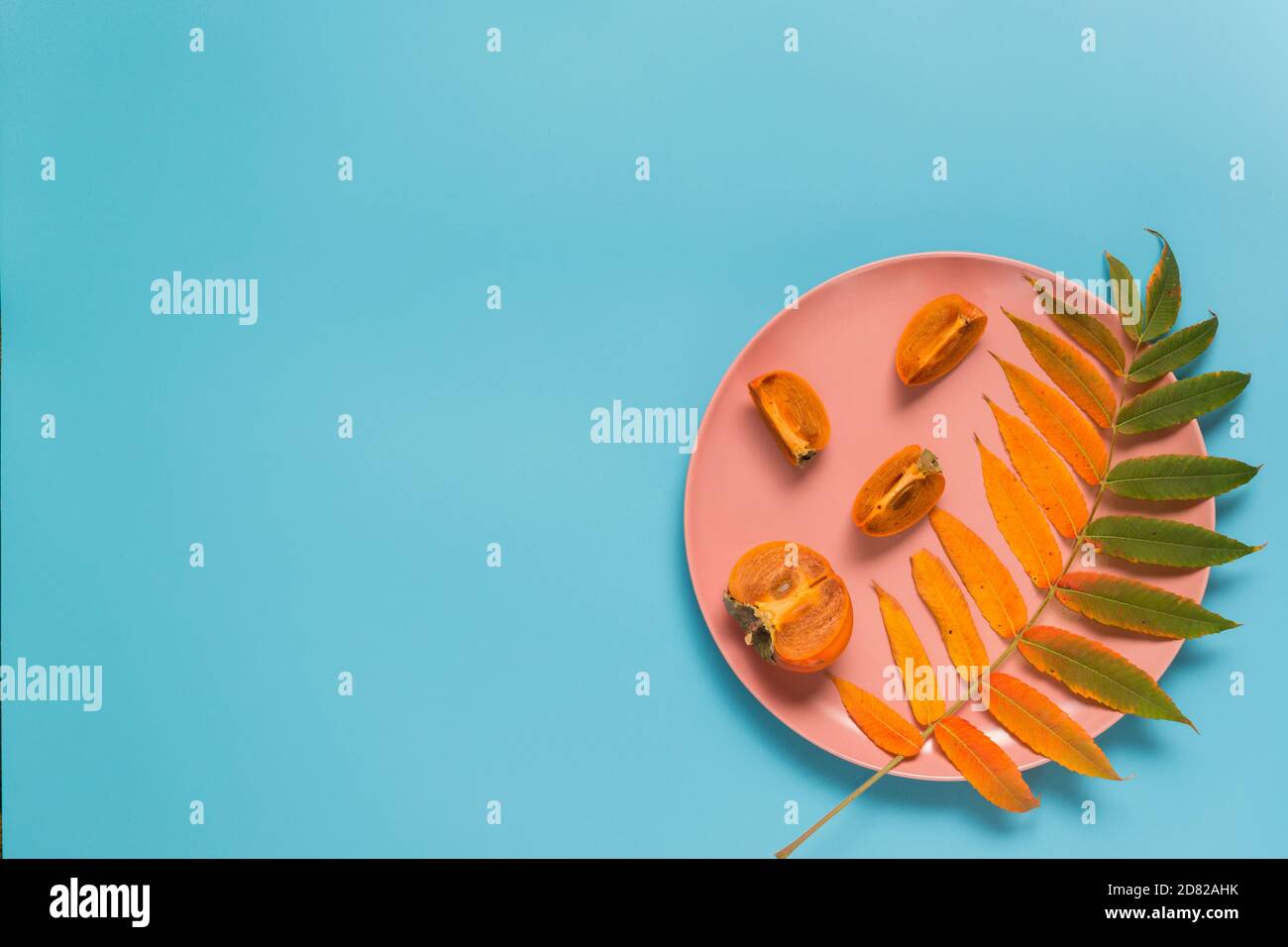 Cartellino di auguri autunnali astratto: Una bella foglia giallastra e un persimmon maturo in un piatto rosa su sfondo blu. Disposizione piatta. Spazio di copia Foto Stock