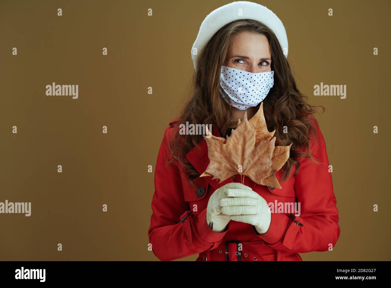 Vita durante la pandemia di covid-19. Ritratto di donna trendy pensiva in camice rosso con foglia di acero giallo autunno isolato su sfondo marrone. Foto Stock