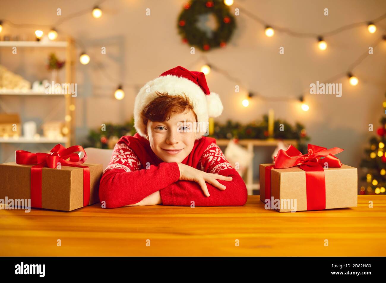 Ragazzo positivo in abiti di Natale guardando la macchina fotografica al tavolo circondato da scatole presenti Foto Stock