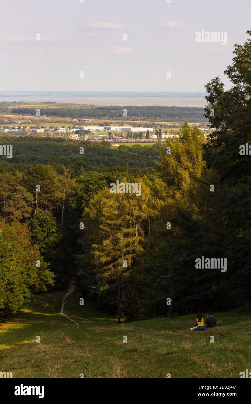 Vista dalla cima della pista di sci al lago fermo con giovane coppia seduta sull'erba, Sopron, Ungheria Foto Stock