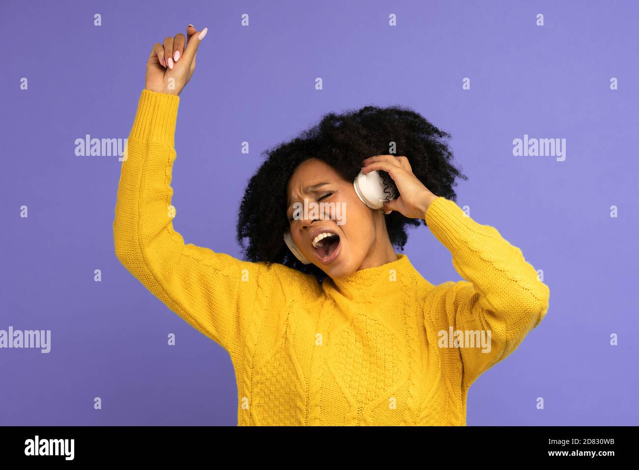 Donna biraciale energizzata che canta, sorride ampiamente, indossa cuffie wireless, ha un buon umore, isolato su sfondo viola. Felice pelle scura riccia Foto Stock