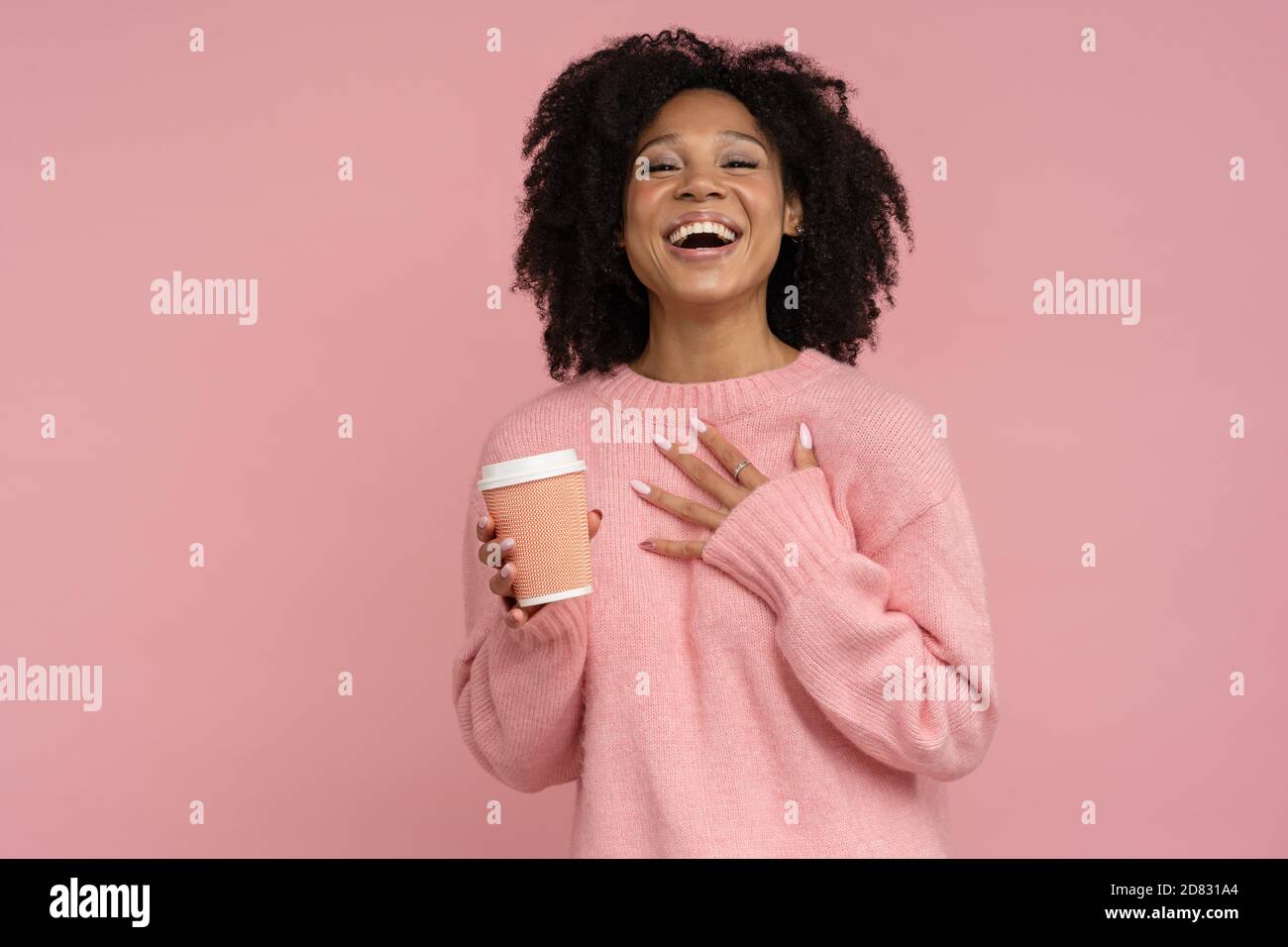 Donna millennial con pelle scura positiva allegra e capelli ricci, indossa un maglione rosa guardando la macchina fotografica, tenendo una tazza di caffè caldo o un tè da asporto. Foto Stock