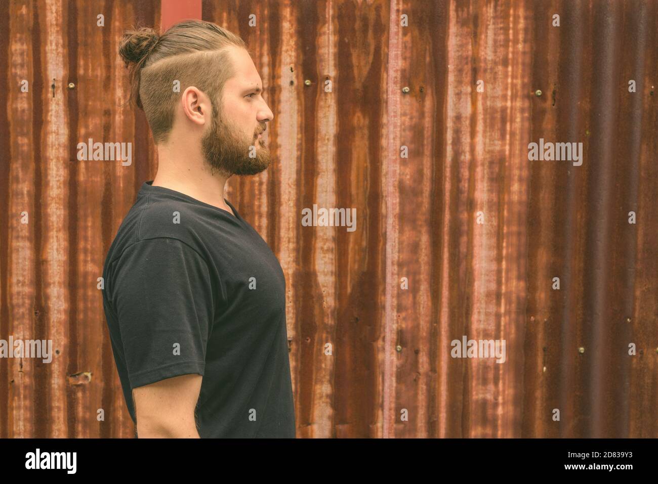 Vista di profilo del giovane uomo barbuto con i capelli legati contro il vecchio arrugginito parete del foglio Foto Stock