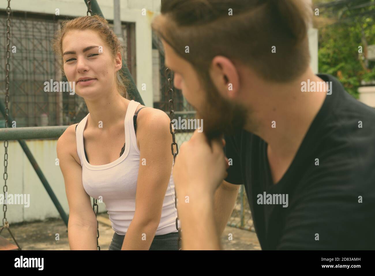 Giovane donna felice guardando il giovane uomo barbuto sorridente e seduta su metallo oscilla insieme nell'amore al vecchio parco giochi Foto Stock