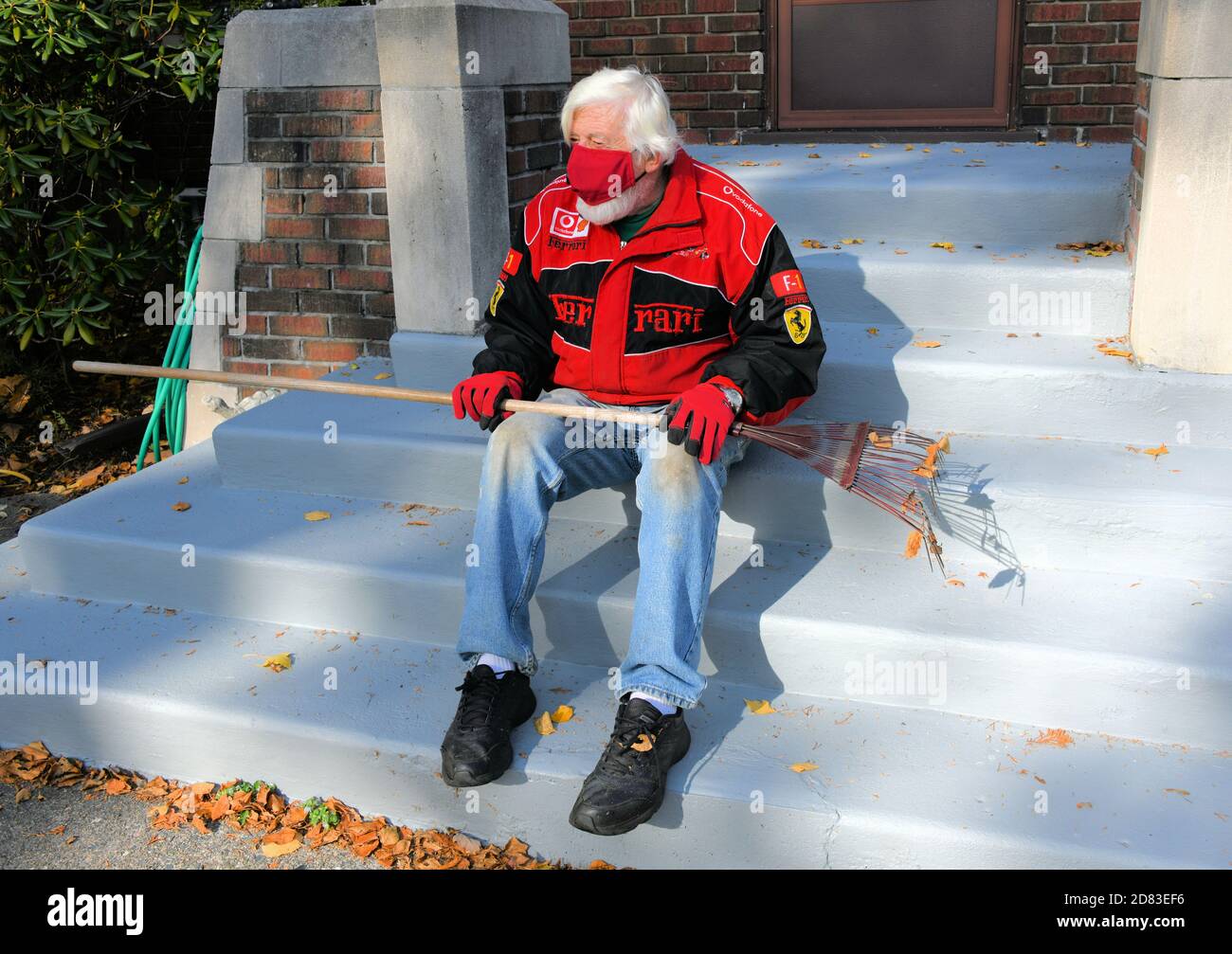 Uomo anziano seduto sulle scale con maschera sopra mentre tiene un rastrello. Foto Stock