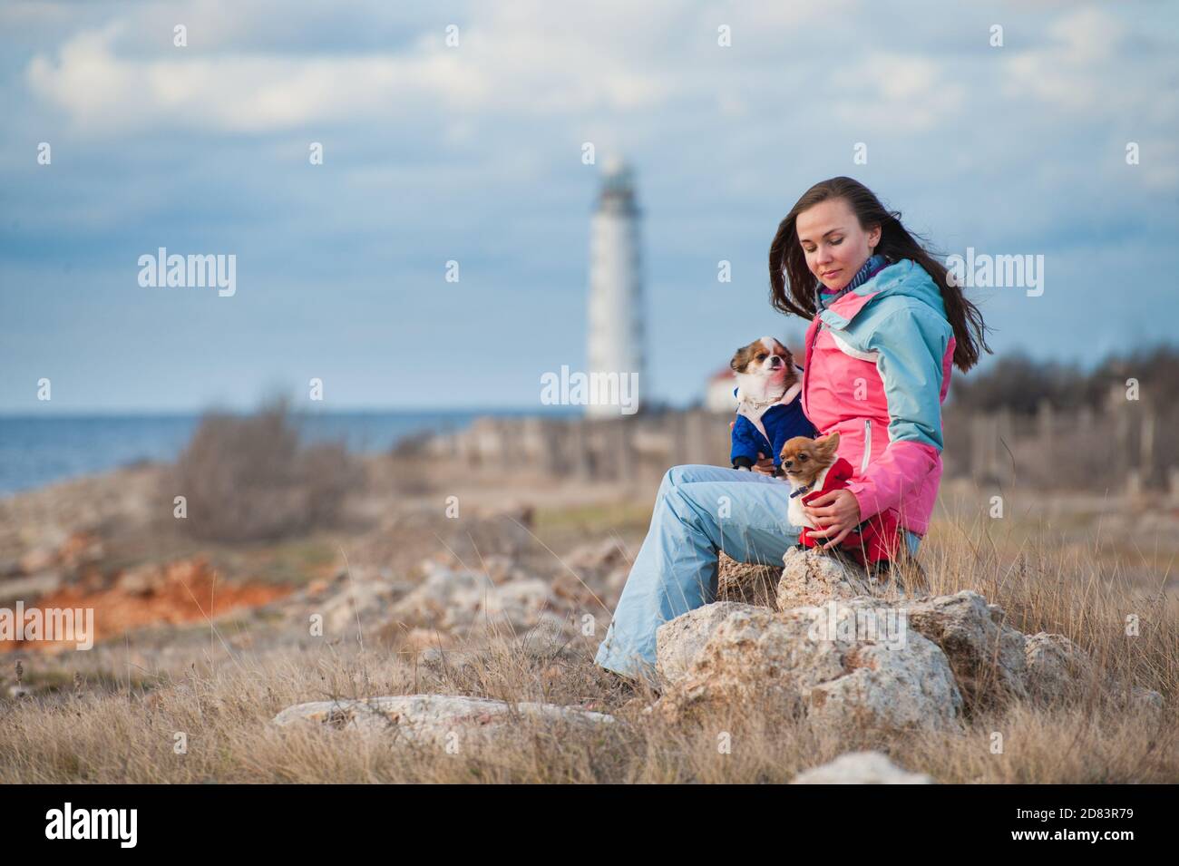 bella giovane donna in tuta in pista seduta su roccia con due piccoli cani da compagnia chihuahua sulla costa invernale autunnale con faro Foto Stock