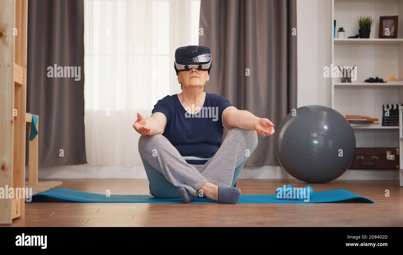 Donna anziana che medita sul tappetino yoga indossando il visore vr. Attivo sano stile di vita sportivo vecchia persona allenamento home wellness ed esercizio interno Foto Stock