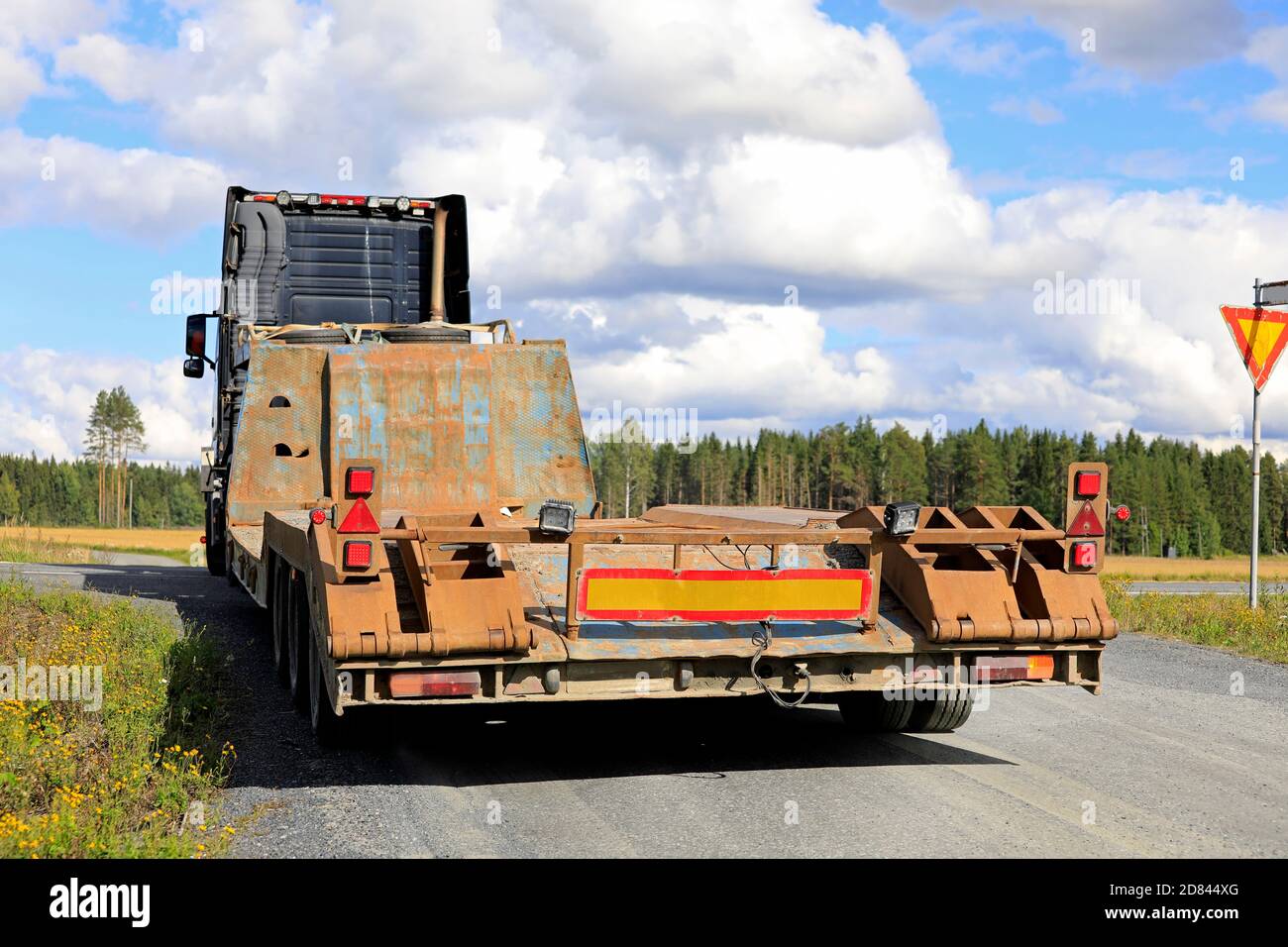 Semi Truck che traina un rimorchio con caricatore basso vuoto su una strada stretta di campagna facendo una svolta a destra verso l'autostrada. Vista posteriore, spazio di copia in alto a destra. Foto Stock