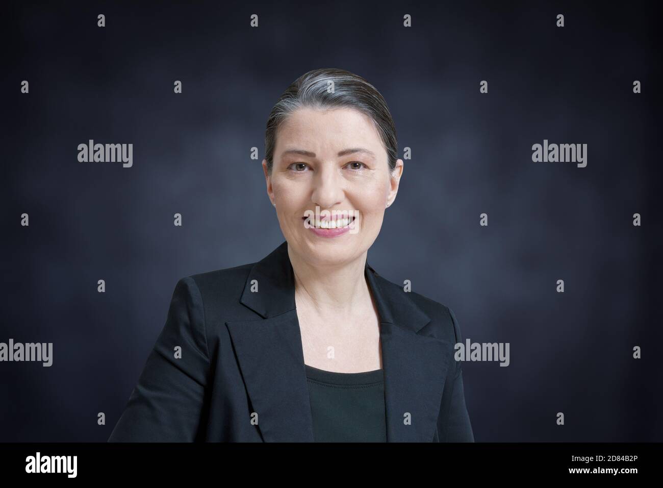 Amichevole sorridente donna di mezza età in un blazer nero su sfondo scuro: professore, avvocato, avvocato o donna d'affari Foto Stock