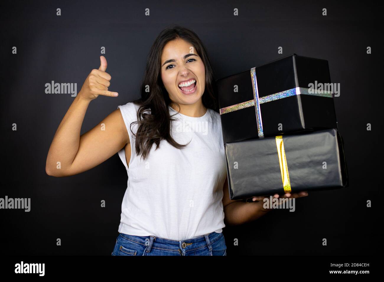 Giovane bella donna che tiene regali su sfondo nero isolato gridando con espressione pazza fare simbolo rock con le mani in su Foto Stock