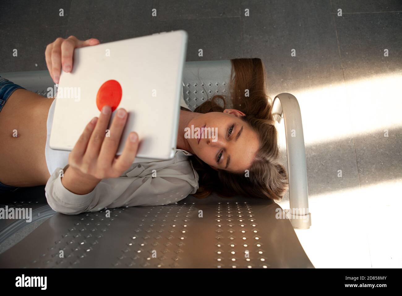 Donna caucasica felice in abiti casual alla moda che tiene tablet e sorridente. Ragazza sicura sdraiata sul sedile in ferro in una sala d'attesa che effettua una videochiamata Foto Stock