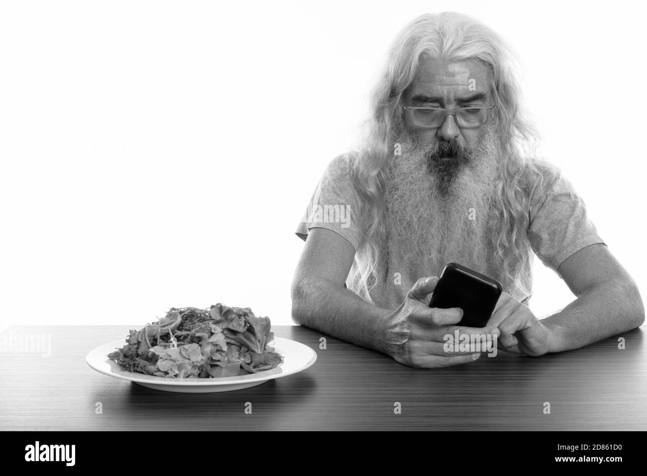 Studio shot di senior uomo barbuto utilizzando il telefono cellulare con piatto di insalata sul tavolo di legno Foto Stock