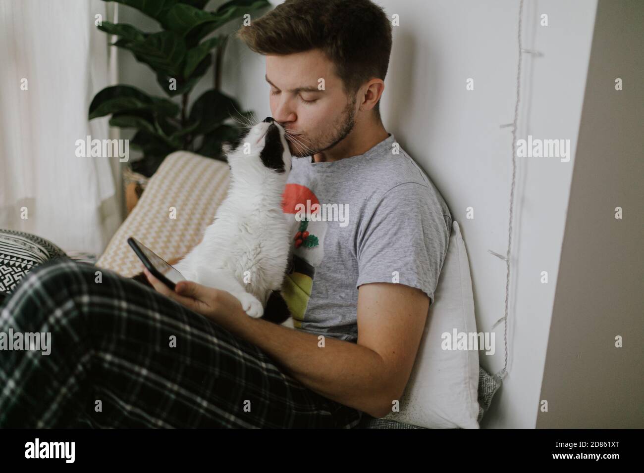 Simpatico gatto che gioca e bacia proprietario e attirare l'attenzione da smartphone. Bell'uomo seduto con il gatto e tenendo il telefono in mano in elegante moderno Foto Stock