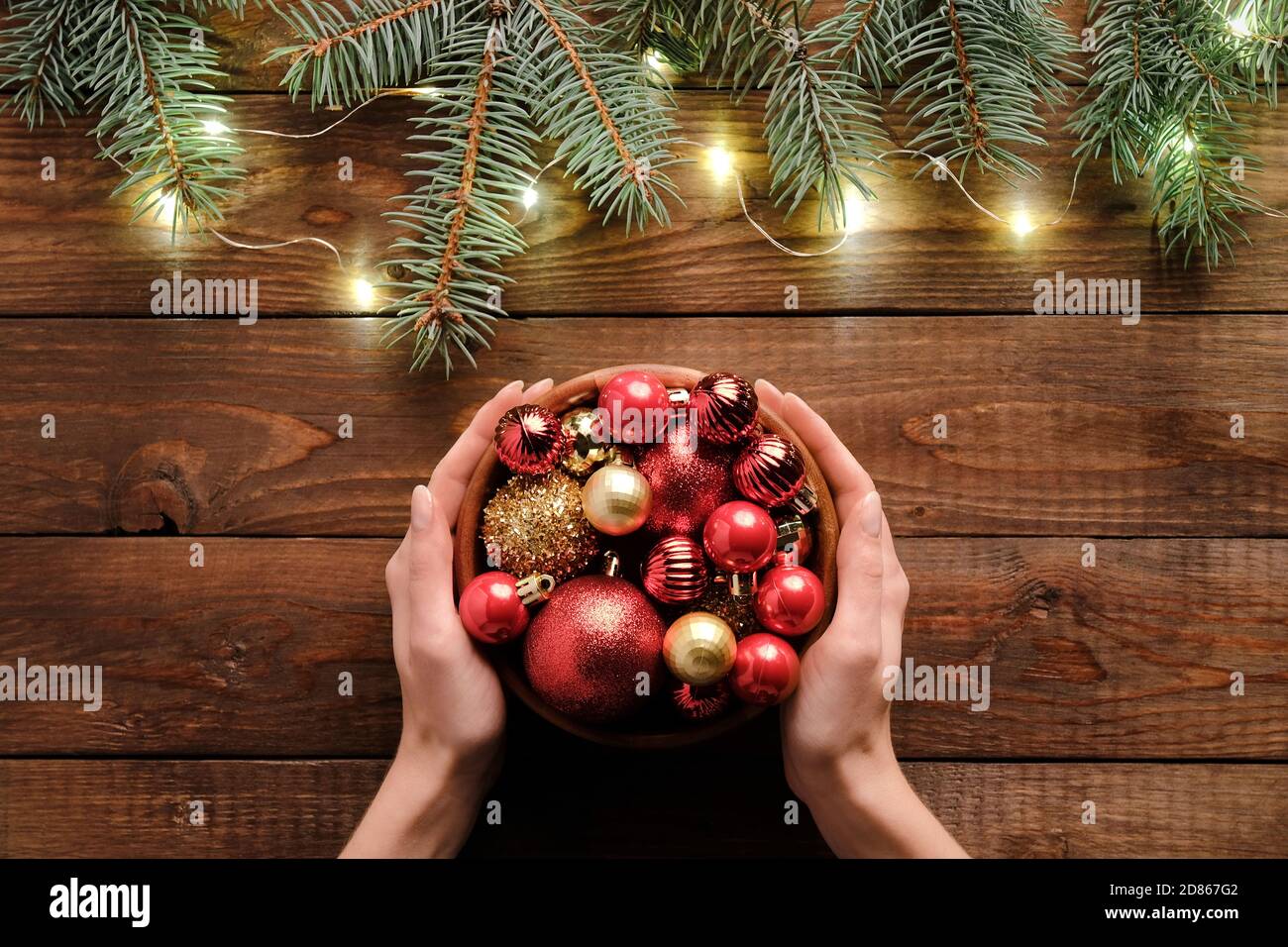 Concetto di preparazione di Natale. Ciotola femminile con decorazione baubles rosso su rustico sfondo di legno con rami di abete e luci. Foto Stock