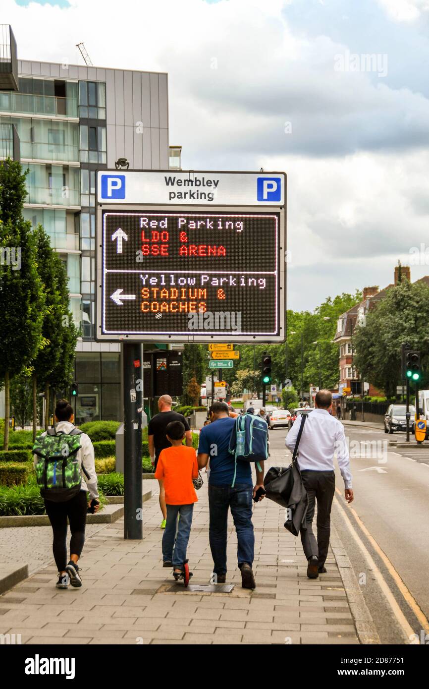 LONDRA, INGHILTERRA - LUGLIO 2018: Persone che camminano lungo una strada sotto un display elettronico con informazioni sul parcheggio del veicolo per lo Stadio di Wembley e l'Arena Foto Stock