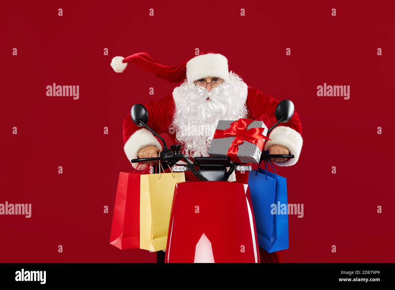 Ritratto di un allegro pazzo Babbo Natale su una moto retrò. Consegna regali su sfondo rosso dello studio. Regali e vendite di Natale e Capodanno Foto Stock