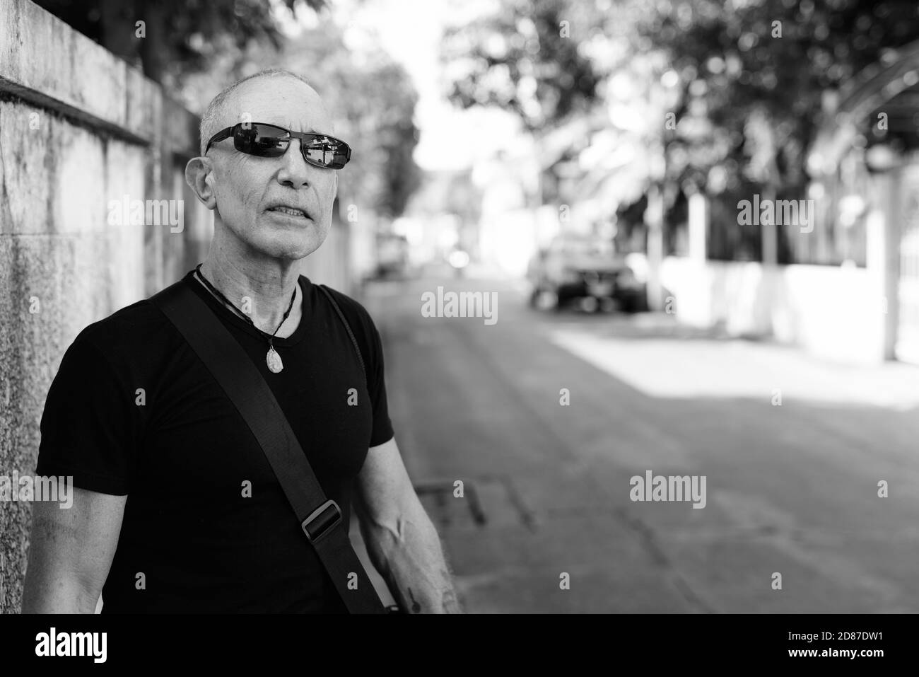 Uomo turistico calvo che pensa mentre si indossano occhiali da sole contro gli anziani muro di cemento nelle strade all'aperto Foto Stock