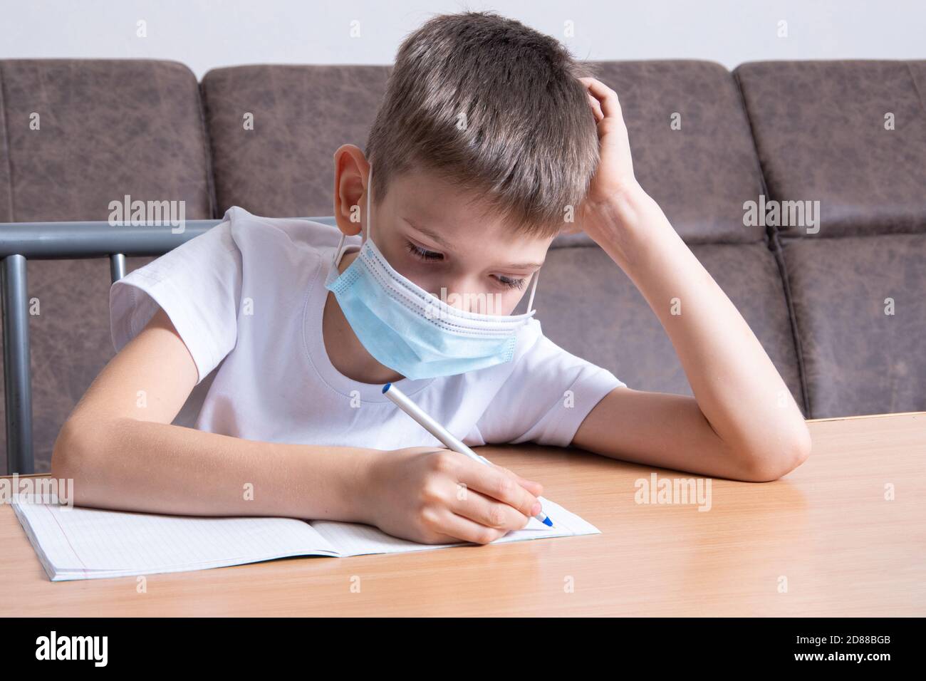 Un ragazzo in una maschera protettiva sul suo viso sta studiando online, scrivendo informazioni in un notebook, seduto alla sua scrivania a casa. Concetto di formazione online, d Foto Stock