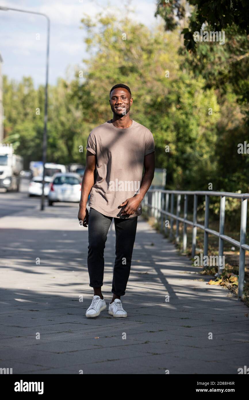 Giovane uomo nero che cammina sul marciapiede della città guardando la macchina fotografica e sorridendo. Lunghezza completa. Verticale. Foto Stock