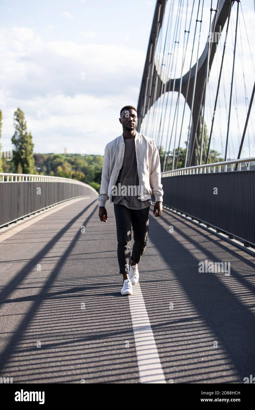 Giovane uomo nero che cammina con un'espressione facciale sicura sul ponte della città. Verticale. Lunghezza completa. Foto Stock