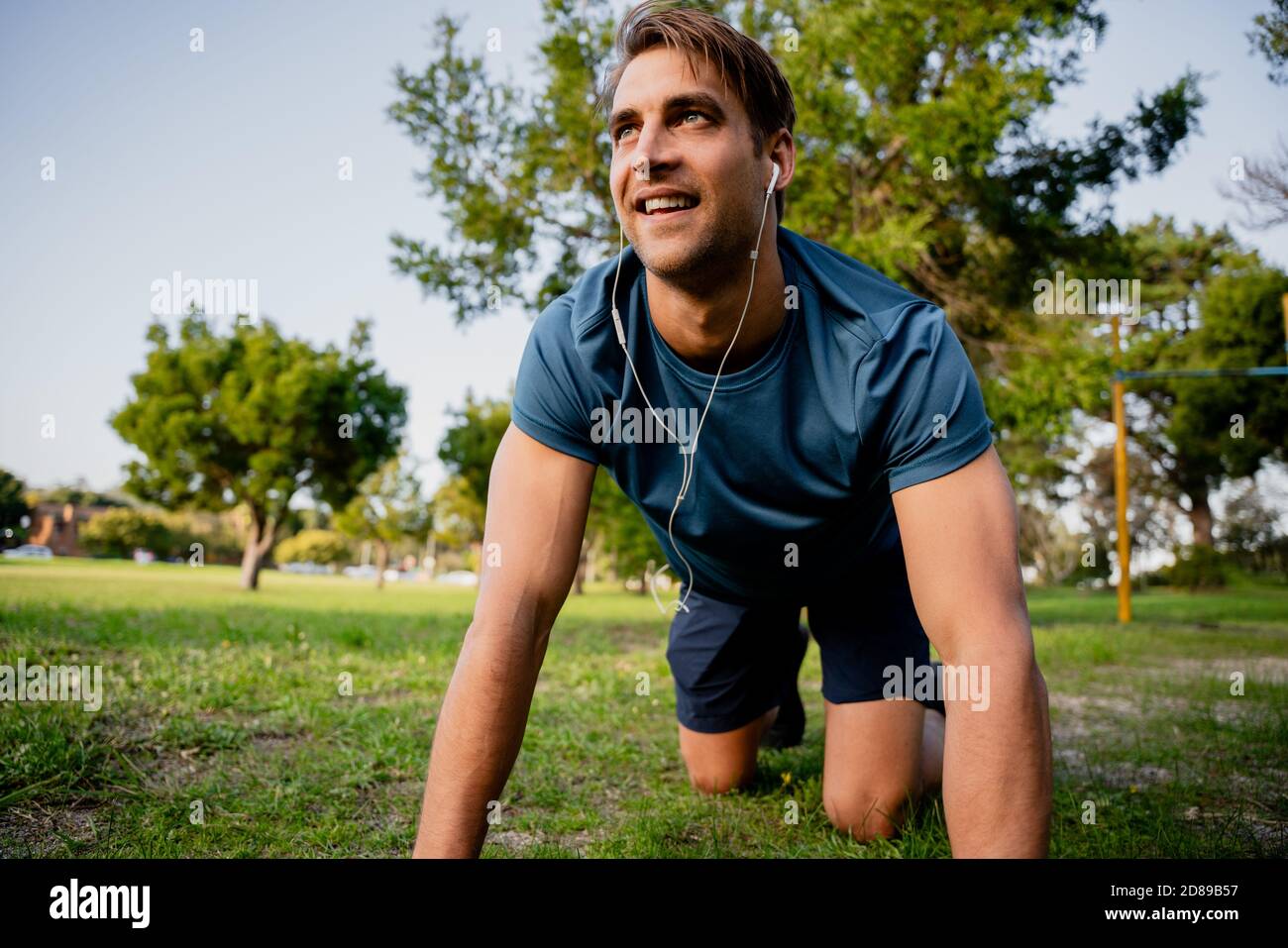 Attraente giovane atleta maschio sorridente sulle mani e sulle ginocchia che si esercitano in un lussureggiante parco all'aperto con auricolari per la musica Foto Stock