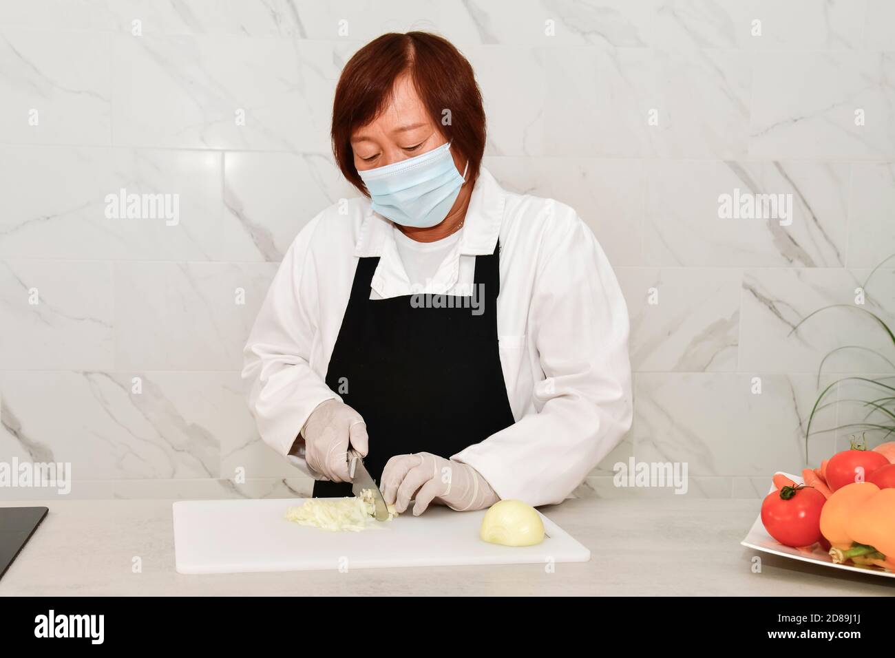 Donna asiatica matura con maschera facciale e guanti che affettano una cipolla pelata su un tagliere in cucina. Concetto di sicurezza e cucina. Foto Stock