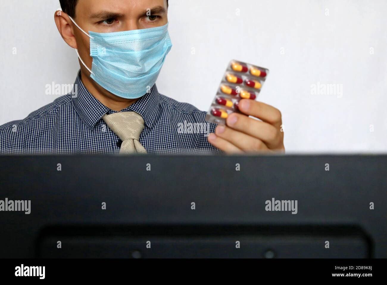 Uomo in maschera di faccia e abiti da ufficio guarda le pillole seduti al monitor del PC. Assunzione di farmaci in capsule, sicurezza sul lavoro durante il coronavirus Foto Stock