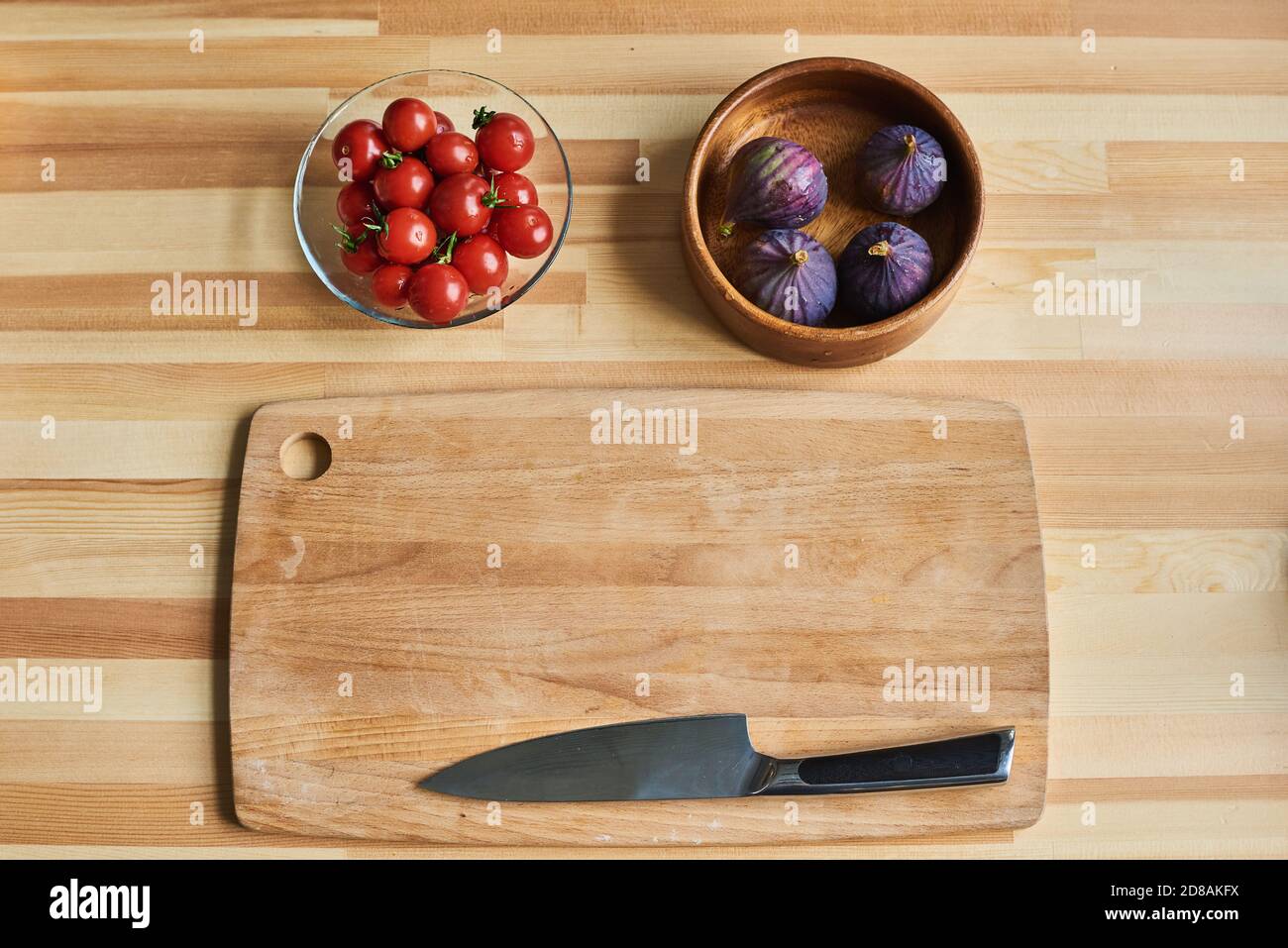 Vista ad angolo alto del tagliere in legno con coltello con pomodori e fichi nelle ciotole sul tavolo Foto Stock