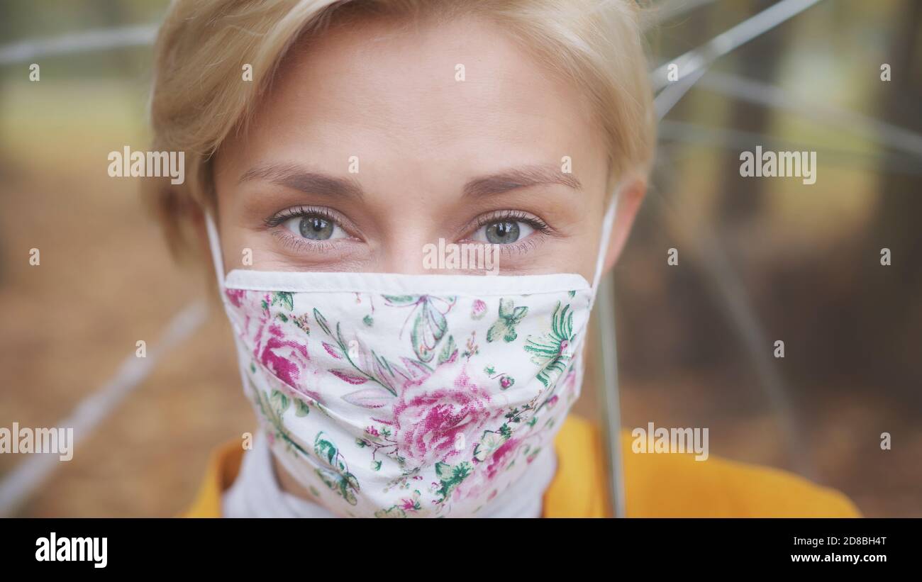 Giovane donna bionda con maschera facciale alla moda e cappotto giallo che tiene ombrello tranparetn nel parco. Foto di alta qualità Foto Stock