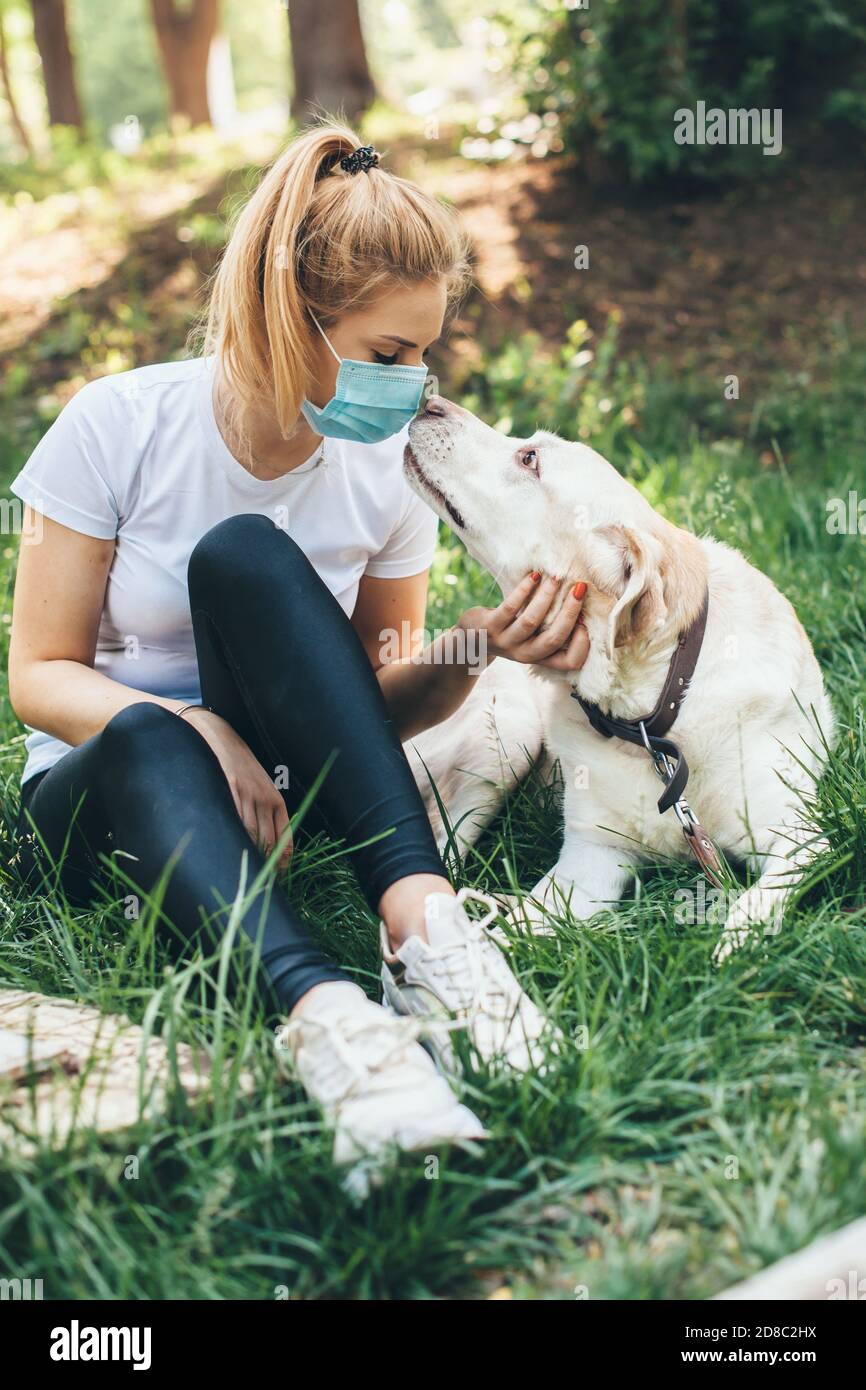 Donna caucasica bionda con maschera sta godendo del tempo all'esterno con il suo cane sdraiato sull'erba in un parco Foto Stock