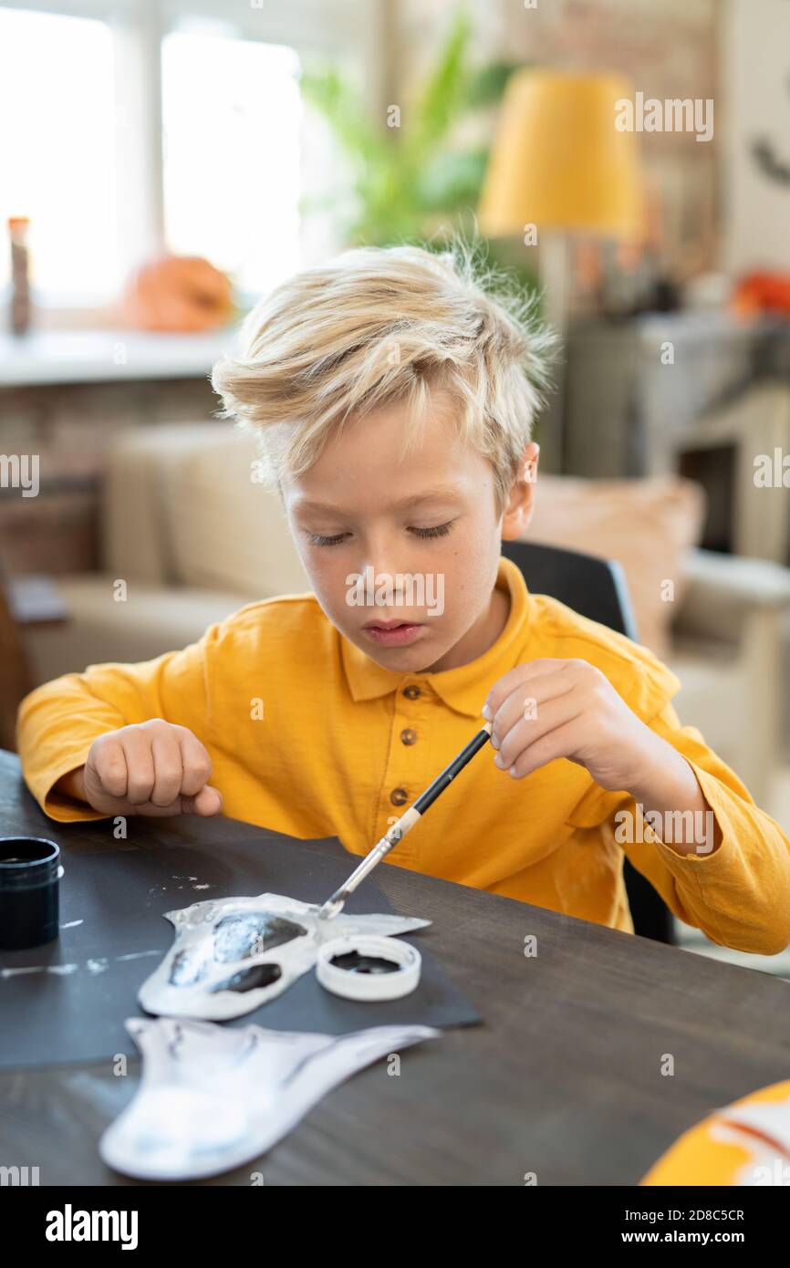 Ragazzo focalizzato con capelli biondi in maglione giallo seduto alla scrivania e. pittura di artigianato fantasma con pennello a classe d'arte Foto Stock