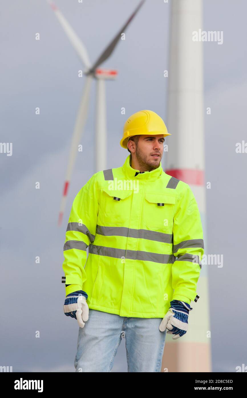 Sorridente giovane ingegnere con elmetto e abbigliamento protettivo davanti alle turbine eoliche sfocate sullo sfondo Foto Stock