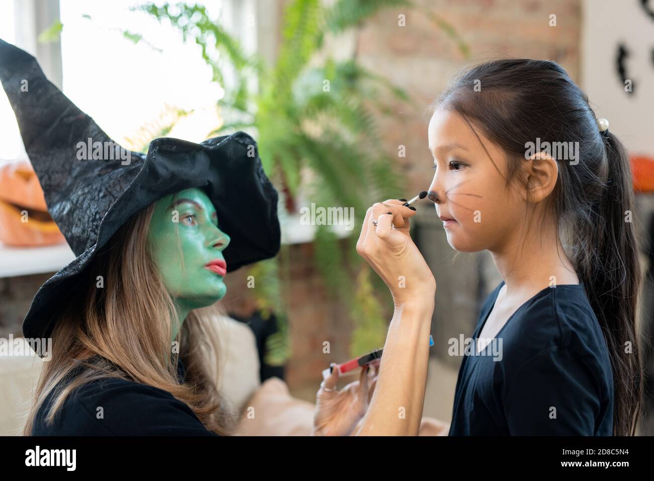 Giovane donna concentrata con painting viso verde whiskers sui bambini Faccia mentre la prepara per la celebrazione di Halloween Foto Stock