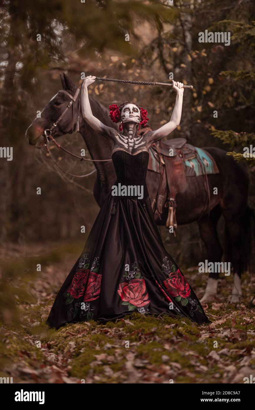 Giovane donna vestita come il simbolo messicano del giorno della calavera morta in un vestito nero che si posa in la foresta con un cavallo Foto Stock