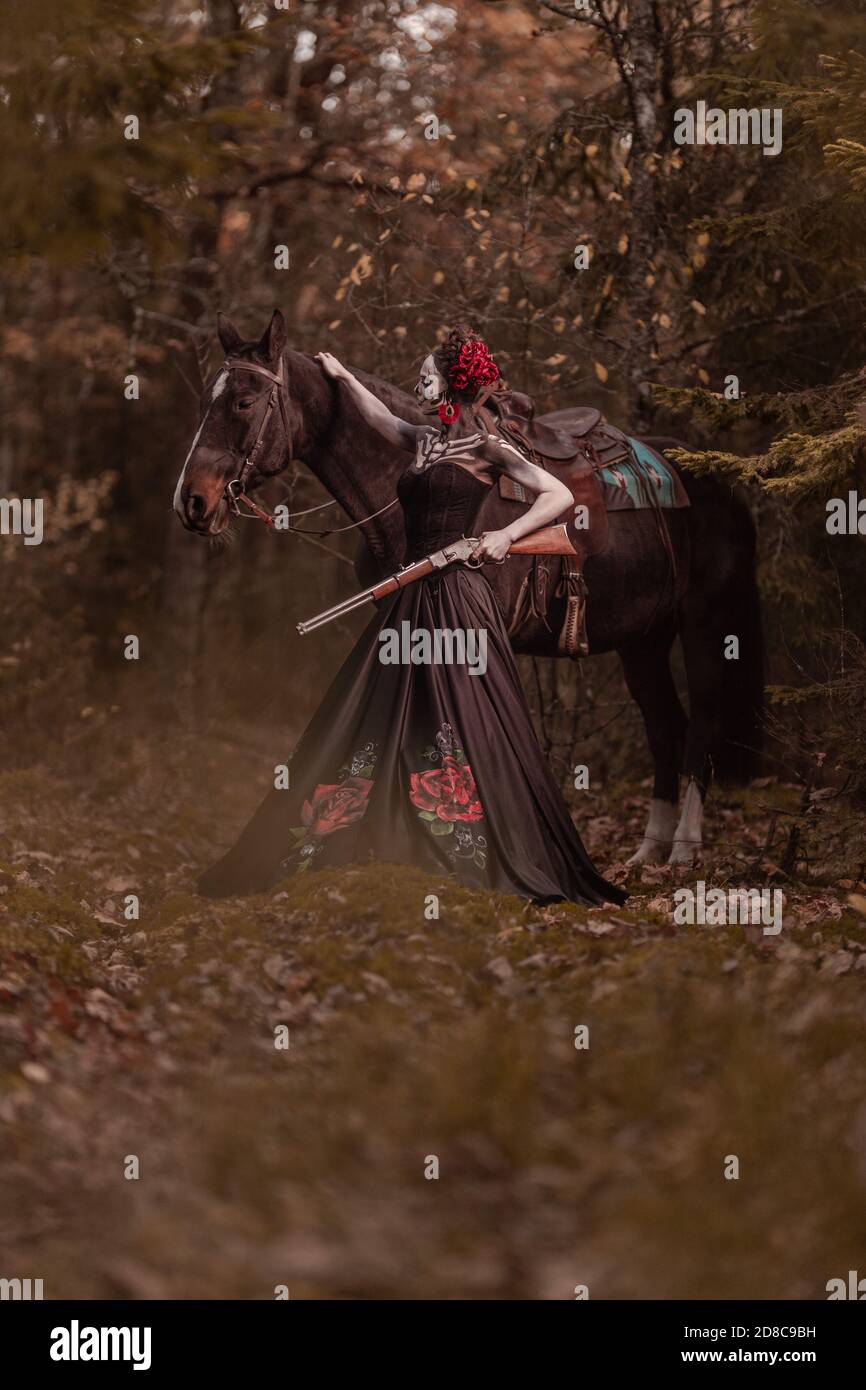 Giovane donna vestita come il simbolo messicano del giorno della calavera morta in un vestito nero che si posa in la foresta con un cavallo Foto Stock